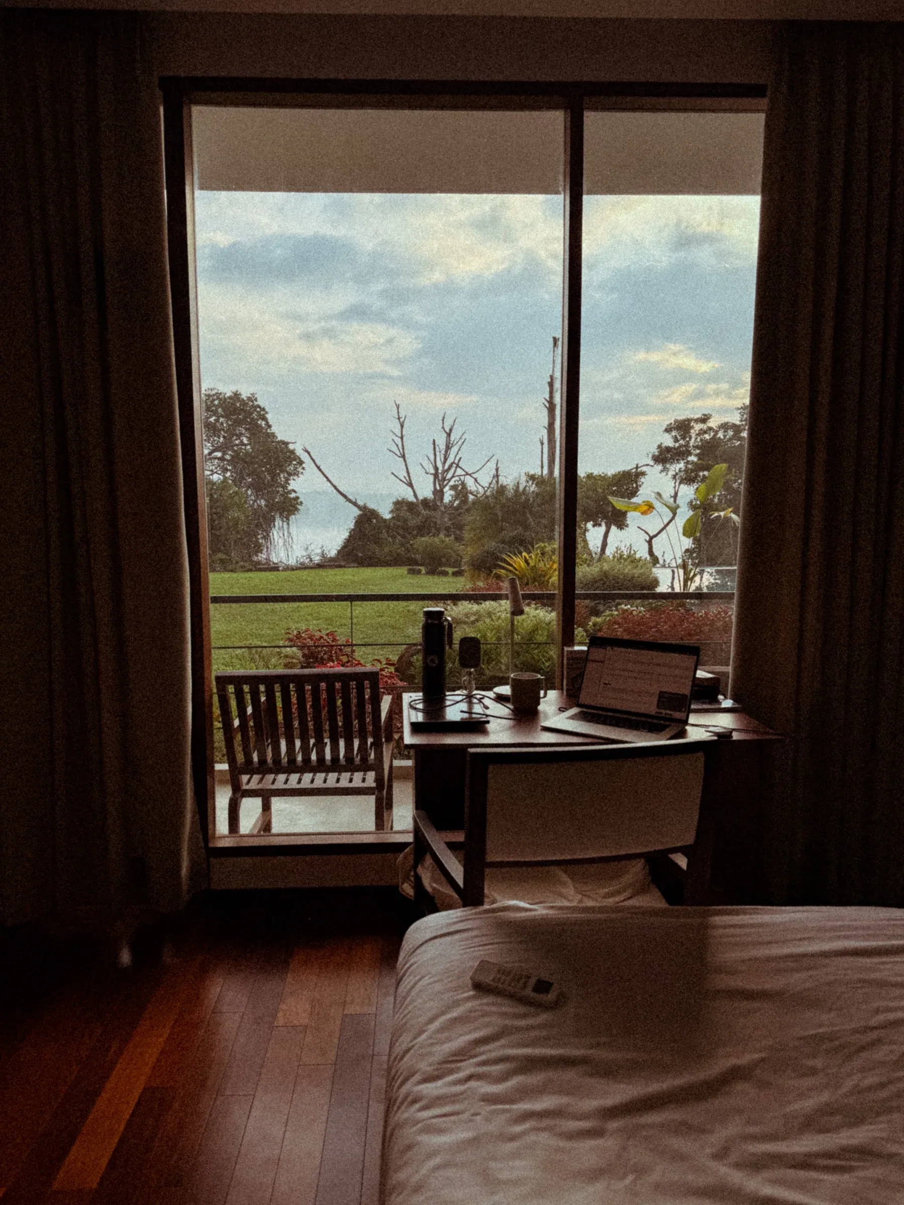 Ein Schreibtisch mit Laptop an einem bodentiefen Fenster, mit Blick über die Wiese auf den See