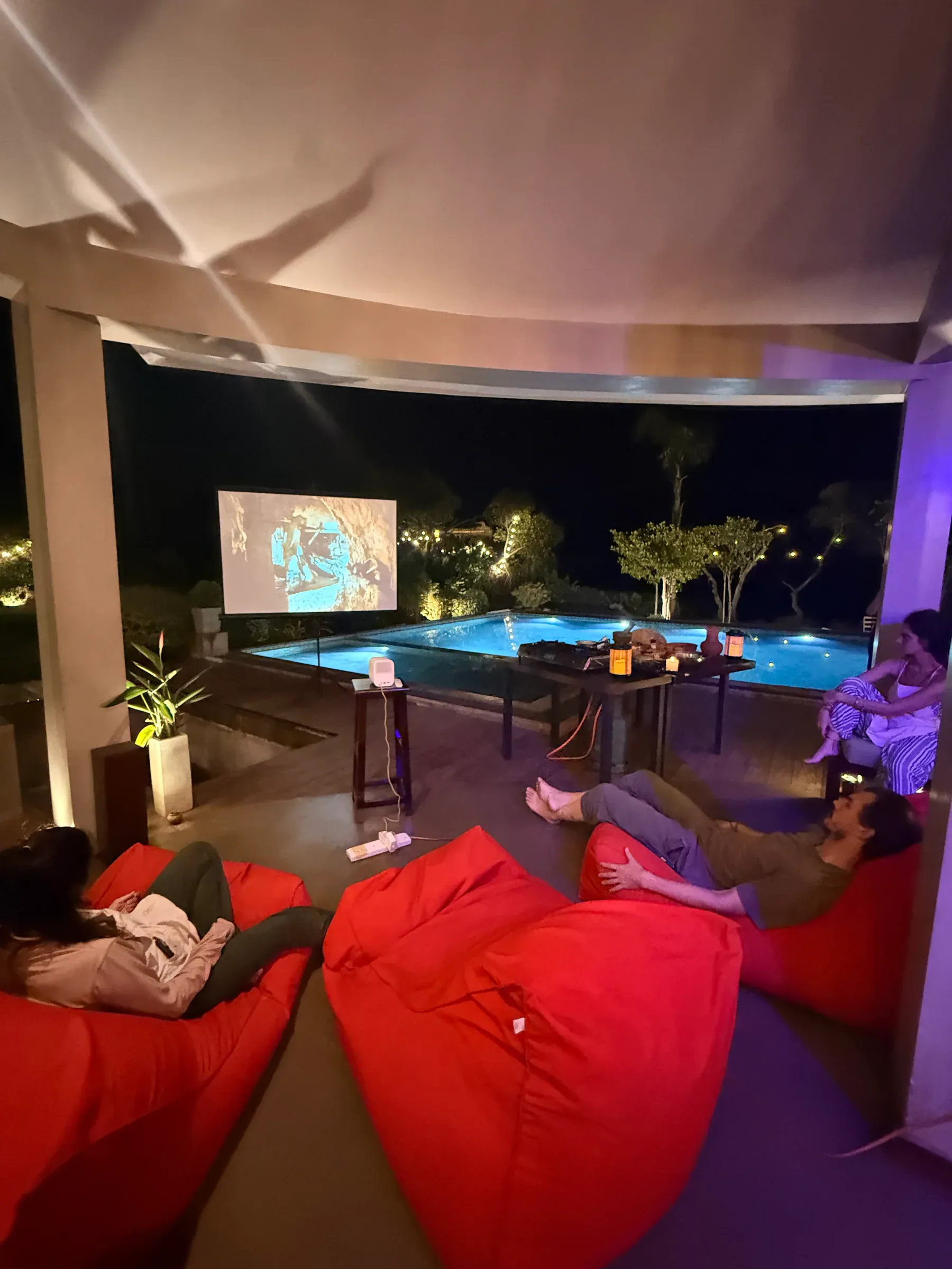 Gäste auf roten Sitzsäcken schauen unter der überdachten Terrasse einen Film, dahinter der beleuchtete Pool