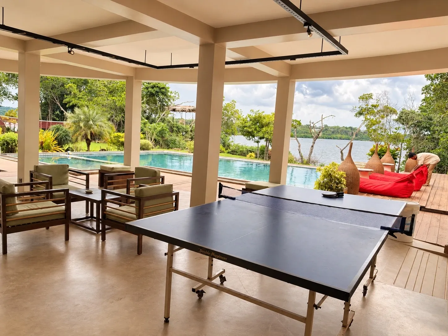 Eine Tischtennisplatte und Sessel in der offenen Lounge neben dem Pool, dahinter der See